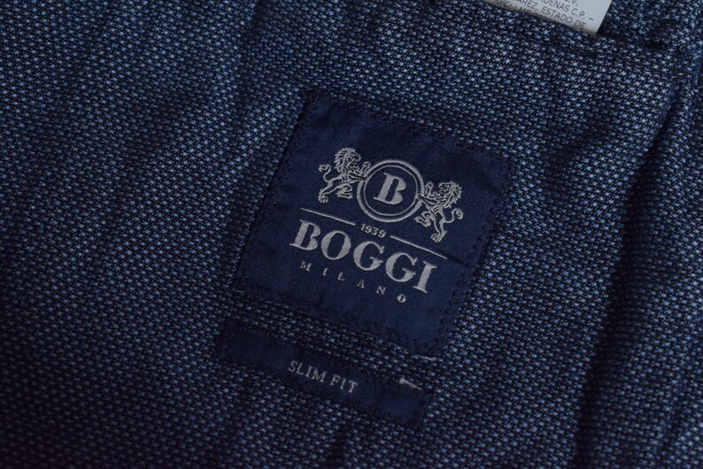 Puede incluir: Primer plano de una tela azul marino con un tejido texturizado. Una etiqueta azul oscuro est&aacute; cosida a la tela, con el nombre de la marca "Boggi Milano" y el texto "Slim Fit". La etiqueta tiene un logotipo con dos leones.