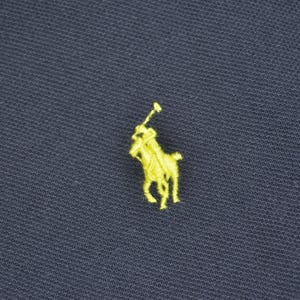 May include: Close-up of navy blue fabric with a small, embroidered yellow logo of a polo player on a horse. The logo is centered and detailed, contrasting with the dark background. The fabric has a textured appearance.