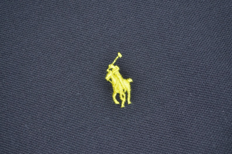May include: Close-up of navy blue fabric with a small, embroidered yellow logo of a polo player on a horse. The logo is centered and detailed, contrasting with the dark background. The fabric has a textured appearance.