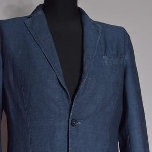 Puede incluir: Un blazer azul marino con un sutil patr&oacute;n texturizado. La chaqueta presenta una solapa con muescas, un bolsillo en el pecho y cierre de dos botones. El blazer se exhibe en un maniqu&iacute; negro.