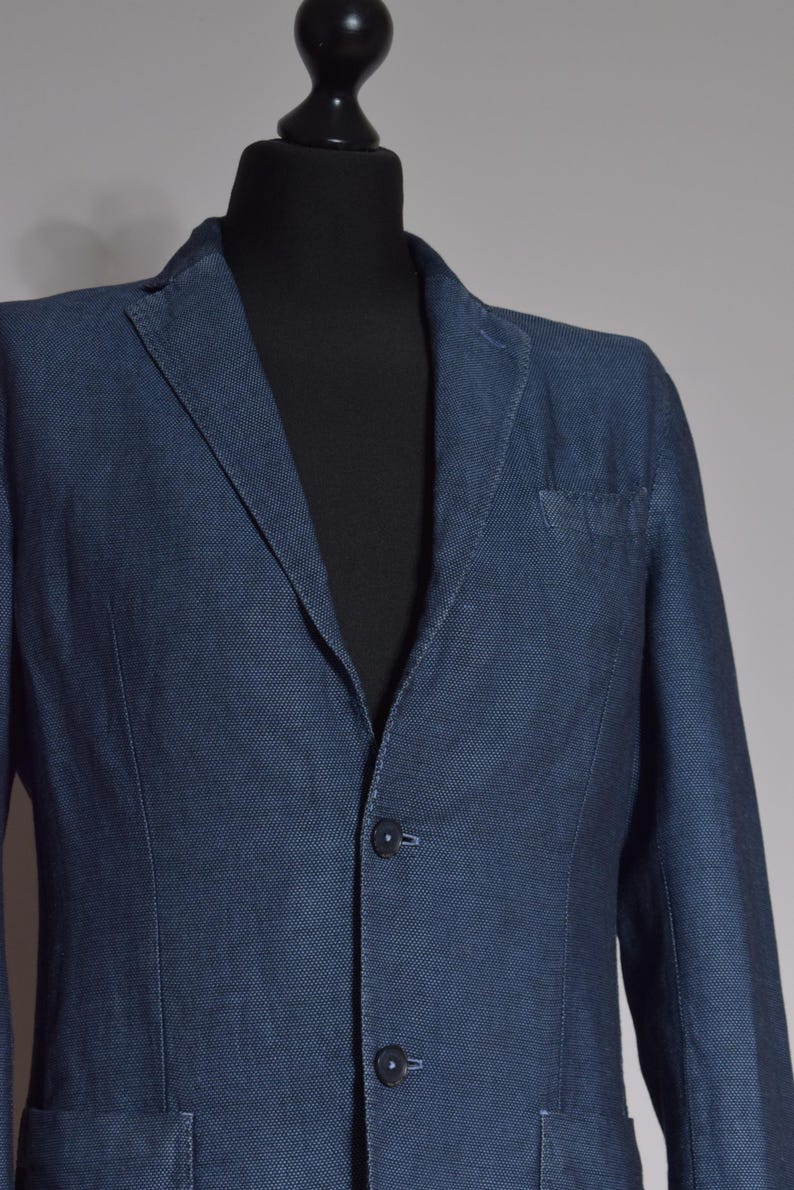 Puede incluir: Un blazer azul marino con un sutil patr&oacute;n texturizado. La chaqueta presenta una solapa con muescas, un bolsillo en el pecho y cierre de dos botones. El blazer se exhibe en un maniqu&iacute; negro.