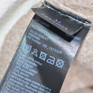 May include: Black care label with white text and symbols. The text includes "ALLSAINTS.COM", "GILDA SWEAT", "WM249M", "PO number: AS_157532R", "50% cotton", "30% polyester", "50% cotton", "20% acrylic", "Machine wash inside out on a delicate cycle, Wash dark colors separately, Reshape while damp, Cool iron on reverse", "Do not dry in dryer".
