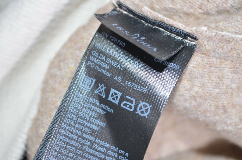 May include: Black care label with white text and symbols. The text includes "ALLSAINTS.COM", "GILDA SWEAT", "WM249M", "PO number: AS_157532R", "50% cotton", "30% polyester", "50% cotton", "20% acrylic", "Machine wash inside out on a delicate cycle, Wash dark colors separately, Reshape while damp, Cool iron on reverse", "Do not dry in dryer".