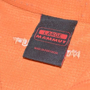 Może przedstawiać: Zbliżenie pomarańczowej tkaniny z czarną etykietą. Na etykiecie widnieje napis "LARGE MAMMUT" w czerwonym tekście, poniżej "MADE IN PORTUGAL". Tkanina ma teksturowany wygląd.