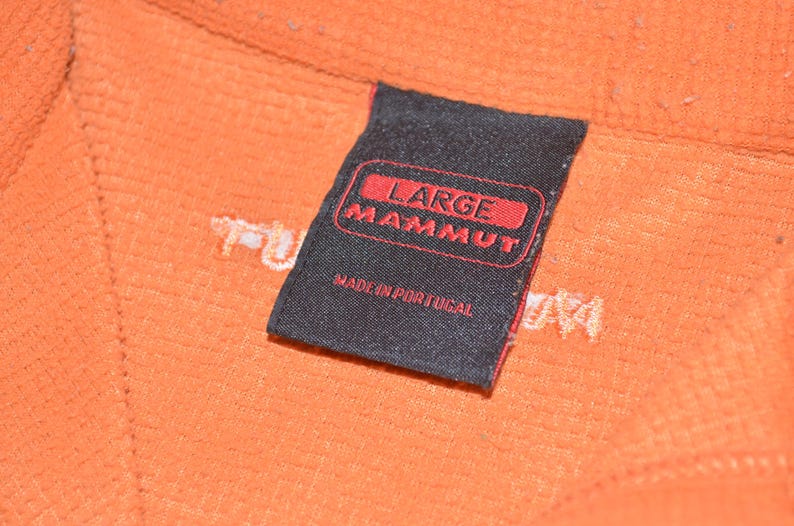 Może przedstawiać: Zbliżenie pomarańczowej tkaniny z czarną etykietą. Na etykiecie widnieje napis "LARGE MAMMUT" w czerwonym tekście, poniżej "MADE IN PORTUGAL". Tkanina ma teksturowany wygląd.