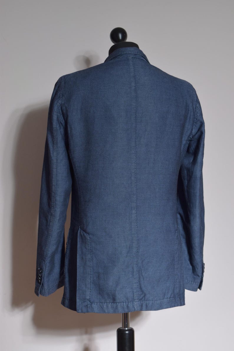 Puede incluir: Una blazer azul marino con cierre de dos botones, bolsillos visibles y solapas con muescas. La chaqueta se exhibe en un maniqu&iacute; negro. La tela tiene un patr&oacute;n sutil.