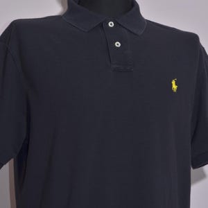 May include: A navy blue polo shirt with short sleeves. The shirt has a classic collar with two buttons and a small yellow logo of a person riding a horse on the left chest.