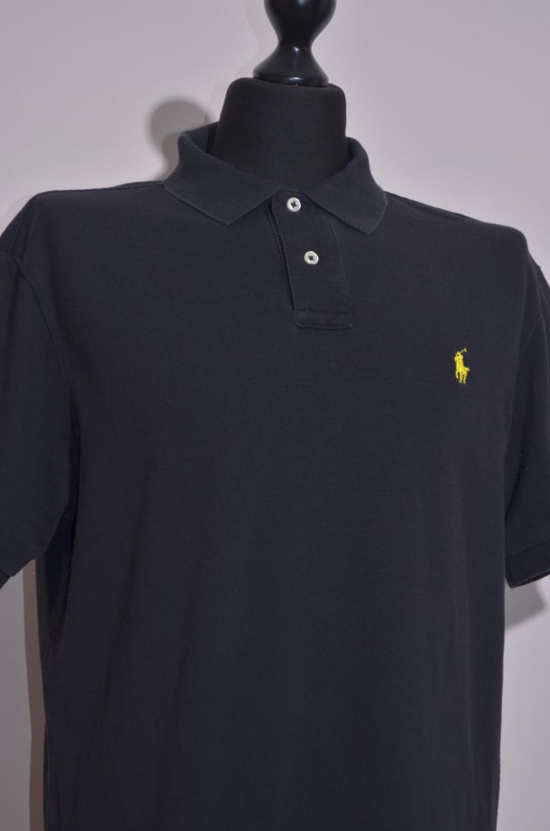 May include: A navy blue polo shirt with short sleeves. The shirt has a classic collar with two buttons and a small yellow logo of a person riding a horse on the left chest.