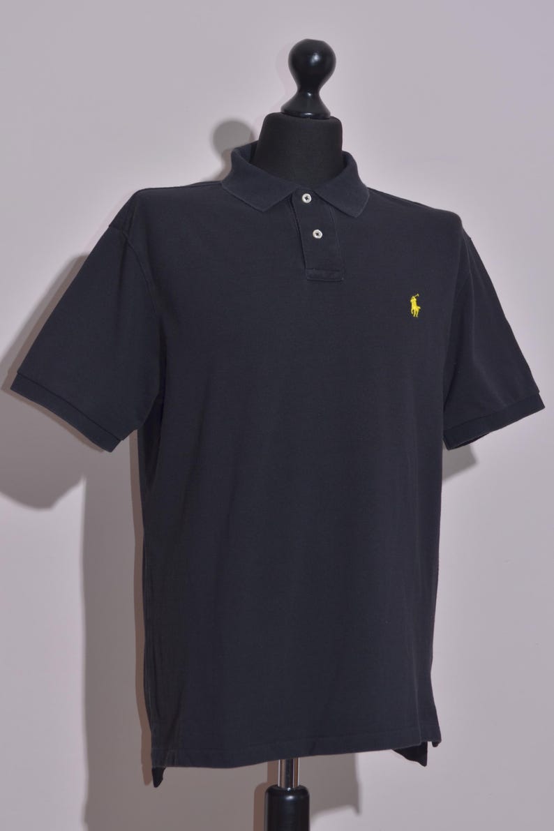 May include: A navy blue polo shirt with short sleeves and a collared neckline. The shirt features a two-button placket and a yellow embroidered logo on the chest. The shirt is displayed on a black mannequin.