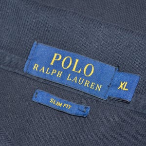 May include: Close-up of a navy blue garment with a blue label featuring the text "POLO RALPH LAUREN" in gold lettering. The label also indicates "XL" size. A smaller label reads "SLIM FIT".