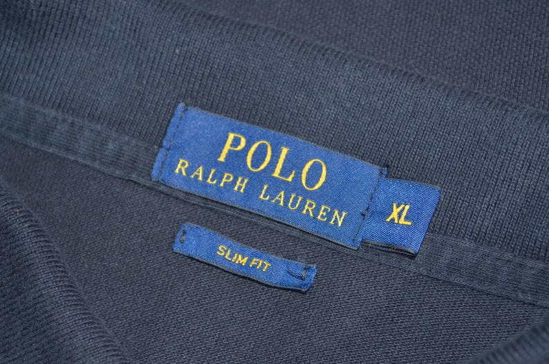 May include: Close-up of a navy blue garment with a blue label featuring the text "POLO RALPH LAUREN" in gold lettering. The label also indicates "XL" size. A smaller label reads "SLIM FIT".