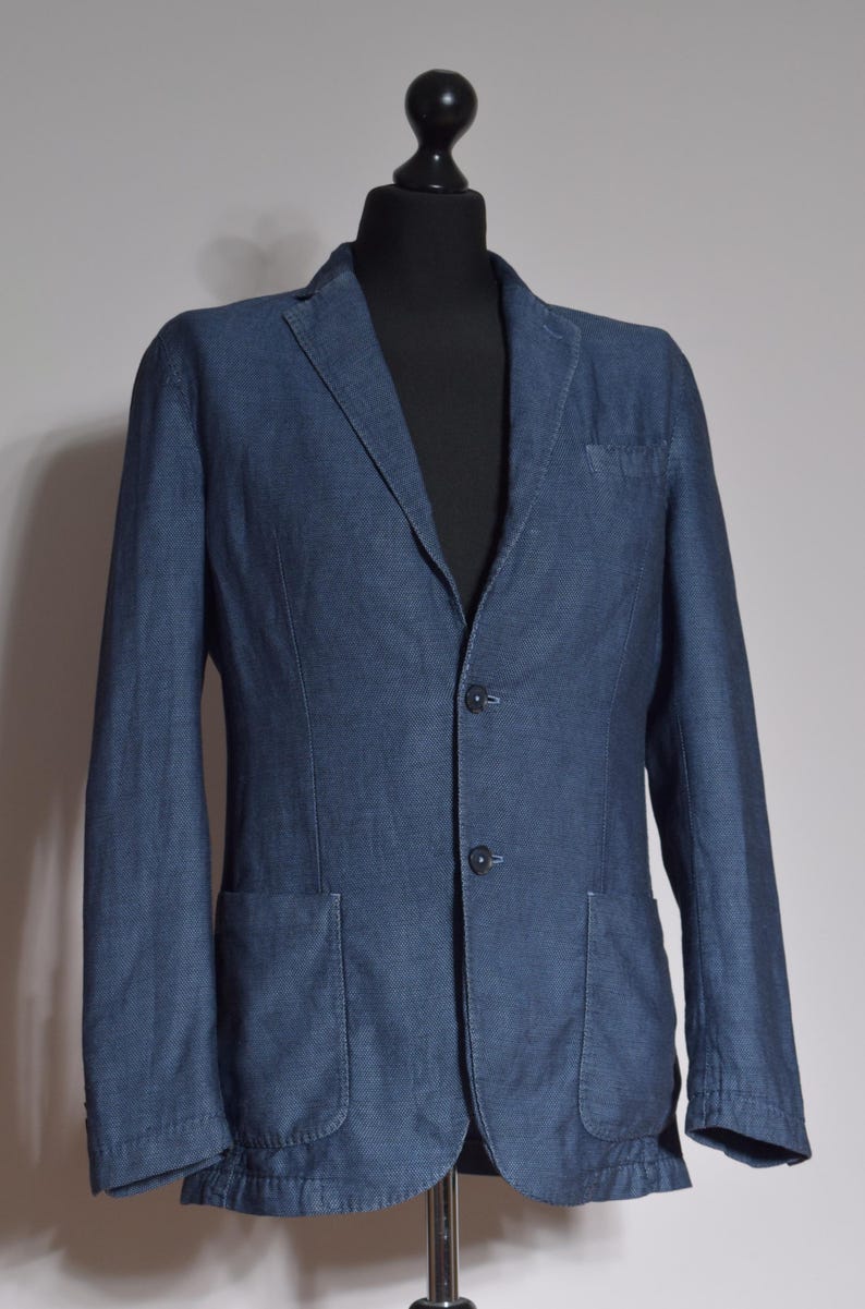 Puede incluir: Un blazer azul con dos bolsillos delanteros, un bolsillo en el pecho y dos botones. El blazer se muestra sobre una cabeza y torso de maniqu&iacute; negros, sobre un fondo neutro. La tela tiene una textura sutil.