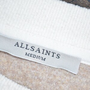May include: A white tag with the text "ALLSAINTS" and "MEDIUM" in black lettering. The tag is attached to a brown and white garment.