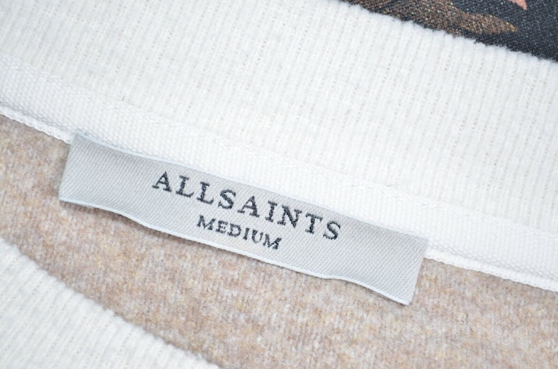 May include: A white tag with the text "ALLSAINTS" and "MEDIUM" in black lettering. The tag is attached to a brown and white garment.