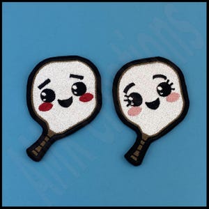 May include: Two white embroidered patches shaped like paddles with smiling faces. The paddles have black eyes, black eyebrows, black mouths, and pink cheeks. The paddles have brown handles.