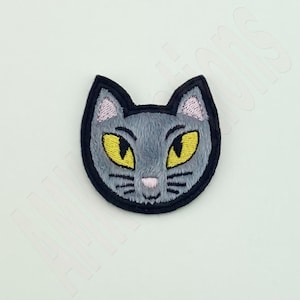 May include: Embroidered patch of a grey cat's face with yellow eyes, a pink nose, and black whiskers. The cat's face is outlined in black, with a fluffy texture. Suitable for application on clothing or accessories.