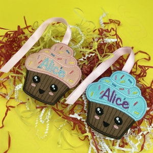 May include: Two pink and blue cupcake-shaped decorations with embroidered faces and the name "Alice" on each. The decorations are hanging from pink and white ribbons against a yellow background.