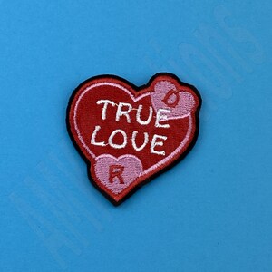 Puede incluir: Parche de fieltro rojo en forma de corazón con bordado blanco que dice "TRUE LOVE". El corazón tiene dos corazones rosas más pequeños en el interior con bordado blanco que dice "R" y "O".