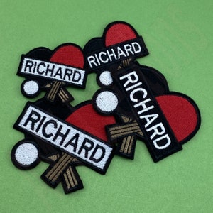 Può includere: Quattro toppe da stirare ricamate in nero e rosso con il nome "RICHARD" in lettere bianche. Le toppe hanno la forma di una racchetta da ping pong e di una pallina.