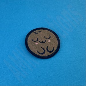 Pode incluir: Um remendo bordado marrom e preto de um coco de desenho animado com um rosto bonito e olhos fechados. O coco tem um contorno preto e está sobre um fundo azul.