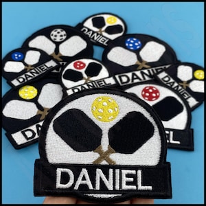 May include: A set of nine embroidered iron-on patches featuring a pair of black pickleball paddles and a yellow pickleball with the name "DANIEL" in white lettering.