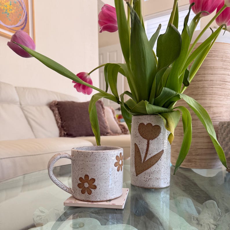 Puede incluir: Una taza de cer&aacute;mica moteada y un jarr&oacute;n cil&iacute;ndrico a juego, ambos con dise&ntilde;os de flores en relieve de color marr&oacute;n. La taza descansa sobre un posavasos cuadrado, mientras que el jarr&oacute;n contiene un ramo de tulipanes rosas sobre cristal.