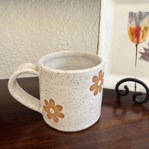 Puede incluir: Una taza de cer&aacute;mica con un esmalte blanquecino moteado y un asa grande y curva. La superficie exterior presenta dos motivos florales pintados a mano en color marr&oacute;n anaranjado, con p&eacute;talos simples y un centro circular.