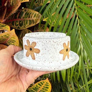May include: A white ceramic yarn bowl with a speckled texture and a wide, flat rim. The exterior features two hand-painted, light brown, six-petaled flowers with dark centers. The bowl has a U-shaped cutout on the top edge for yarn.