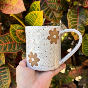 Puede incluir: Una taza de cer&aacute;mica blanca con textura moteada y dos dise&ntilde;os de flores marrones. La taza presenta un cuerpo cil&iacute;ndrico y un asa curva en el lado derecho. La superficie de la taza est&aacute; cubierta con peque&ntilde;as motas marrones sobre la base.