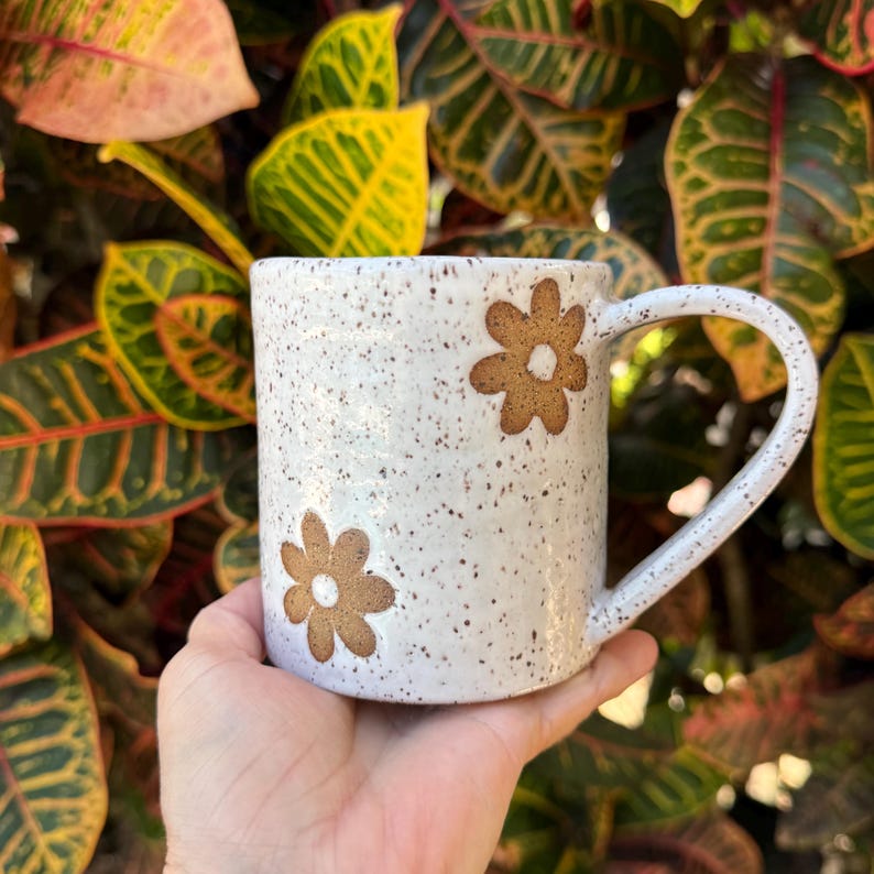 Puede incluir: Una taza de cer&aacute;mica blanca con textura moteada y dos dise&ntilde;os de flores marrones. La taza presenta un cuerpo cil&iacute;ndrico y un asa curva en el lado derecho. La superficie de la taza est&aacute; cubierta con peque&ntilde;as motas marrones sobre la base.