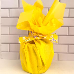 Puede incluir: Un objeto cil&iacute;ndrico envuelto en papel de seda amarillo brillante, asegurado en la parte superior con una cinta amarilla que presenta un patr&oacute;n repetitivo de flores blancas y moradas similares a margaritas. El papel est&aacute; atado y recogido.