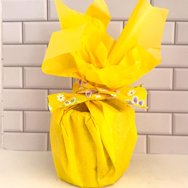 Puede incluir: Un objeto cil&iacute;ndrico envuelto en papel de seda amarillo brillante, asegurado en la parte superior con una cinta amarilla que presenta un patr&oacute;n repetitivo de flores blancas y moradas similares a margaritas. El papel est&aacute; atado y recogido.