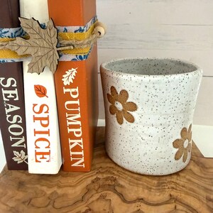 Puede incluir: Una taza de cer&aacute;mica blanca moteada con patrones de flores marrones se encuentra junto a tres libros apilados. Los libros marrones, blancos y naranjas tienen el texto SEASON, SPICE y PUMPKIN, atados con cordel y un recorte de hoja de arce de madera.
