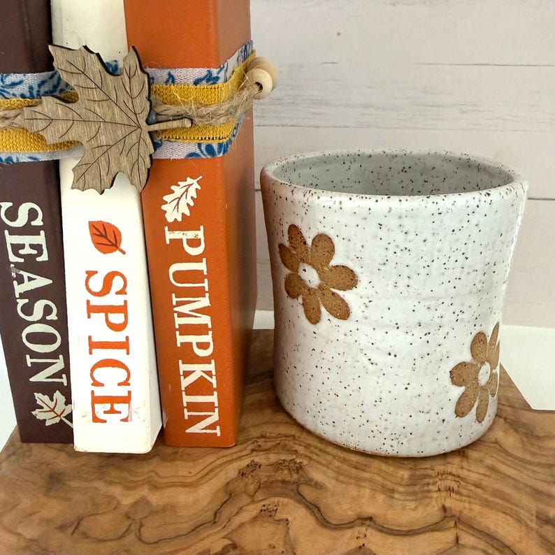 Puede incluir: Una taza de cer&aacute;mica blanca moteada con patrones de flores marrones se encuentra junto a tres libros apilados. Los libros marrones, blancos y naranjas tienen el texto SEASON, SPICE y PUMPKIN, atados con cordel y un recorte de hoja de arce de madera.