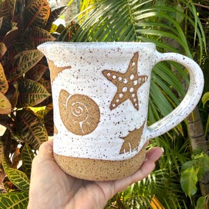May include: A white ceramic pitcher with a speckled finish and a textured brown base. The exterior features embossed brown designs of a starfish, a spiral seashell, and a smaller shell. It includes a curved handle and a small pouring spout.