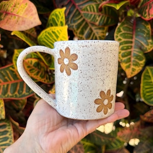 Puede incluir: Una taza de cer&aacute;mica blanca moteada con dos patrones florales marrones en forma de margarita. La taza tiene un cuerpo cil&iacute;ndrico y un asa curva en el lado izquierdo. La superficie tiene un acabado brillante con peque&ntilde;as motas marrones.