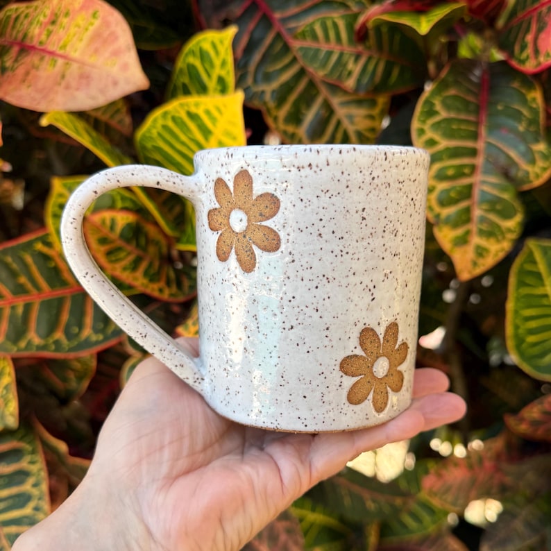 Puede incluir: Una taza de cer&aacute;mica blanca moteada con dos patrones florales marrones en forma de margarita. La taza tiene un cuerpo cil&iacute;ndrico y un asa curva en el lado izquierdo. La superficie tiene un acabado brillante con peque&ntilde;as motas marrones.