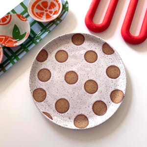 May include: A round ceramic plate with a speckled white glaze and brown polka dots. Beside it sits a rectangular tray featuring a green and blue plaid pattern with orange citrus slice motifs, along with two bright red U-shaped silicone trivets.