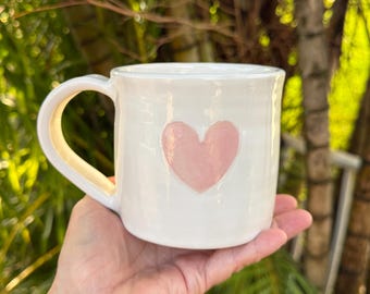 May include: A white ceramic mug with a glossy finish featuring a single, centered, light pink heart design. The mug has a rounded handle on the left side and a smooth, cylindrical body with subtle horizontal ridges along the exterior surface.