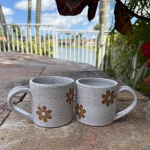 Puede incluir: Dos tazas de cer&aacute;mica con esmalte blanco roto moteado y motivos florales pintados a mano en color marr&oacute;n. Cada taza presenta un cuerpo cil&iacute;ndrico, un asa lateral resistente y un borde liso. La textura moteada es uniforme en el exterior.