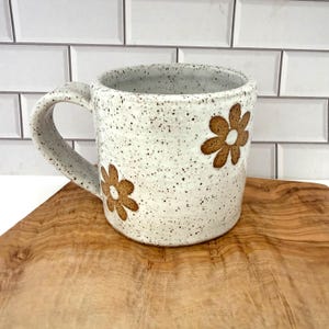 Puede incluir: Una taza de cer&aacute;mica con esmalte blanco moteado y un asa grande y curva. La superficie exterior presenta dos dise&ntilde;os de flores marrones con seis p&eacute;talos cada una. La taza tiene una forma cil&iacute;ndrica y un acabado suave y moteado.
