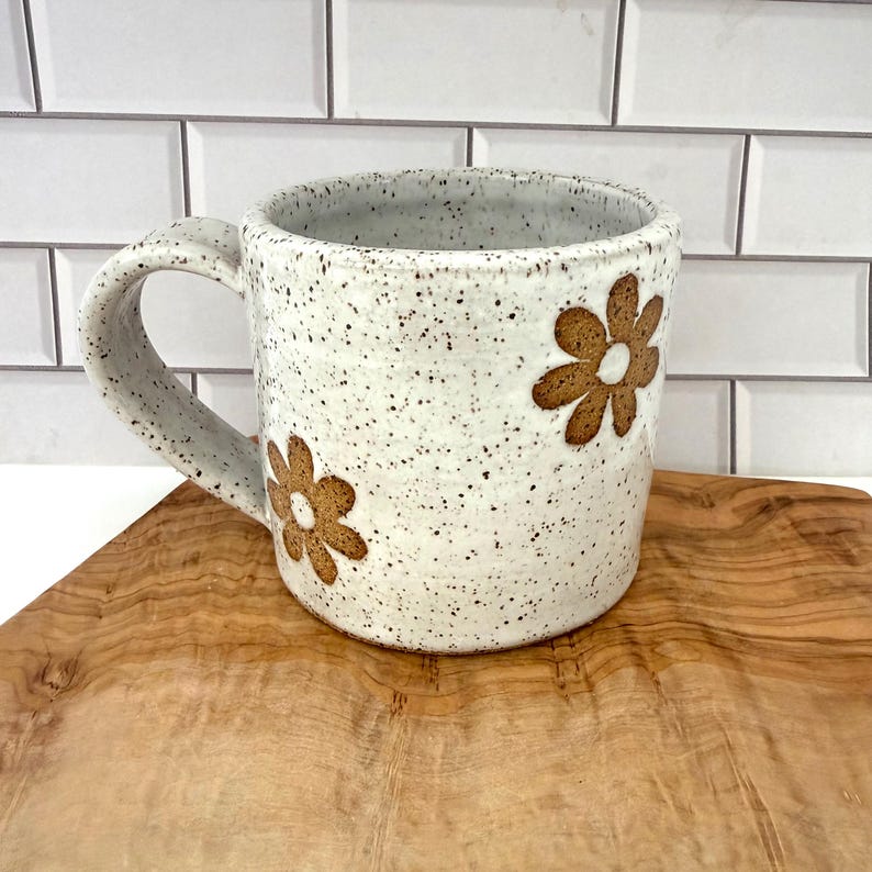 Puede incluir: Una taza de cer&aacute;mica con esmalte blanco moteado y un asa grande y curva. La superficie exterior presenta dos dise&ntilde;os de flores marrones con seis p&eacute;talos cada una. La taza tiene una forma cil&iacute;ndrica y un acabado suave y moteado.