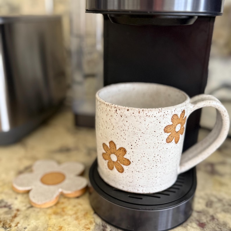 Puede incluir: Una taza de cer&aacute;mica de color blanco roto con motas, decorada con motivos florales marrones y un posavasos a juego en forma de flor. La taza tiene un asa resistente y se apoya sobre la base de una cafetera negra, junto al posavasos.