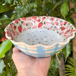 May include: A ceramic colander bowl with a scalloped rim, featuring a white interior with blue speckles and drainage holes. The exterior displays a light blue and cream checkered pattern, while the inner rim is decorated with red strawberries.