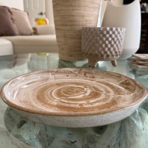 May include: A round ceramic plate featuring a speckled off-white base with a swirling, hand-painted tan and brown glaze pattern on the interior surface. The plate has a low profile with a slightly raised rim and a smooth, glossy finish.