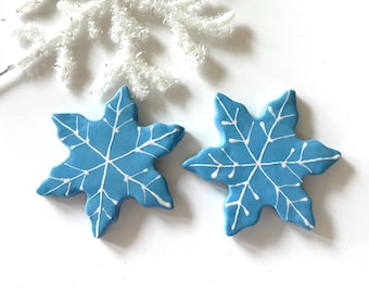 Puede incluir: Dos adornos azules en forma de copo de nieve con detalles blancos. Los adornos tienen un color base azul claro y líneas blancas que crean un diseño de copo de nieve. Una decoración de copo de nieve blanca y brillante está en el fondo.