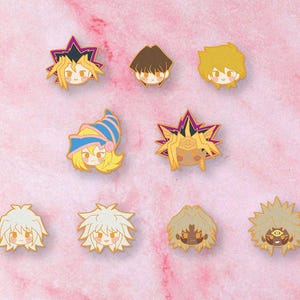 Peut inclure: Collection d'épingles en émail représentant différents personnages avec des accents jaunes, marron et violets. Les épingles sont disposées sur une surface marbrée rose et blanche. Les épingles semblent inspirées d'une série animée populaire.