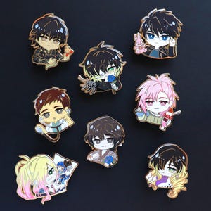 May include: Nine enamel pins showcasing anime-style characters with diverse hairstyles and attire. The pins have gold-coloured metal borders and depict characters holding items such as a rabbit, a flower, and a dessert. The characters have varied hair colours, including black, brown, and pink.