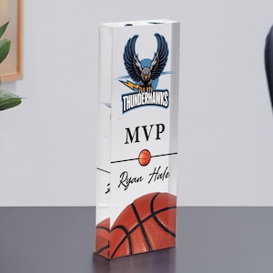 May include: A clear acrylic basketball trophy with the text "Thunderhawks MVP" and a basketball graphic. The name "Ryan Hale" is also printed on the trophy. The trophy is rectangular and stands on a black surface.