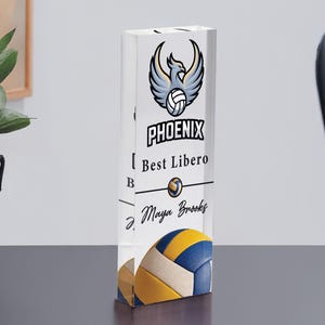 May include: A rectangular volleyball trophy with the text "PHOENIX" and a phoenix logo. The trophy also includes the text "Best Libero" and a volleyball design in blue, yellow, and white. The name "Maya Brooks" is also on the trophy.